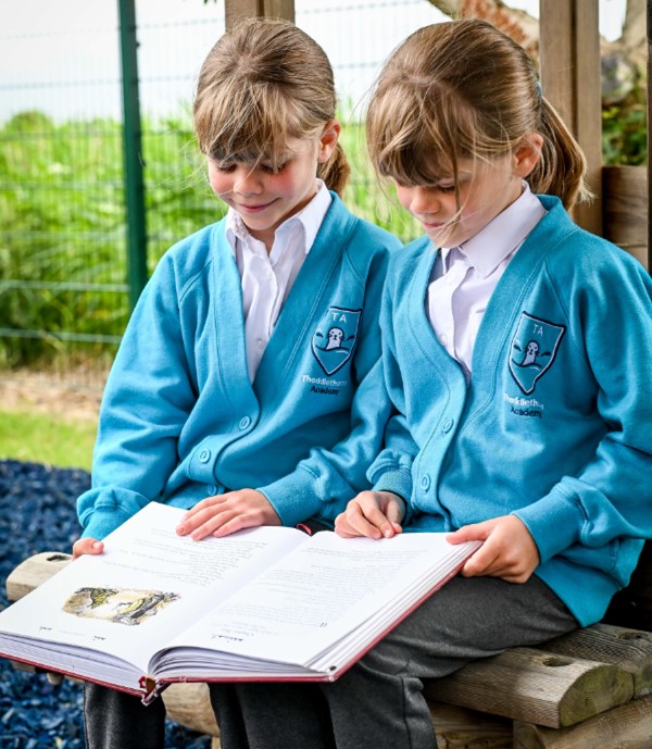 Two Theddlethorpe Academy pupils sitting on a bench outside reading a book.